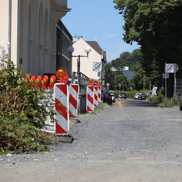 Ein Schotterweg, links stehen rot-weiße Baken, rechts im Hintergrund parkt ein Auto.