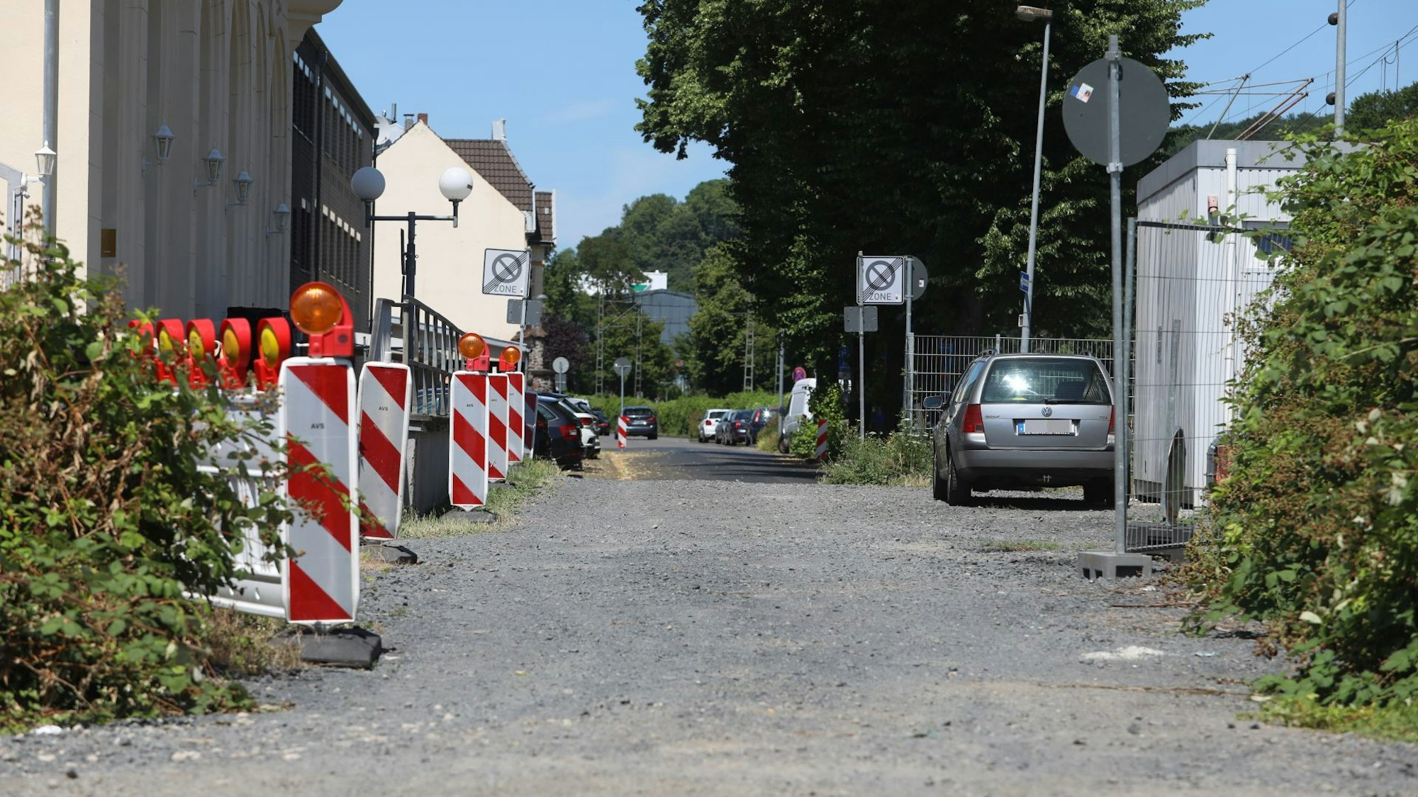 Ein Schotterweg, links stehen rot-weiße Baken, rechts im Hintergrund parkt ein Auto.