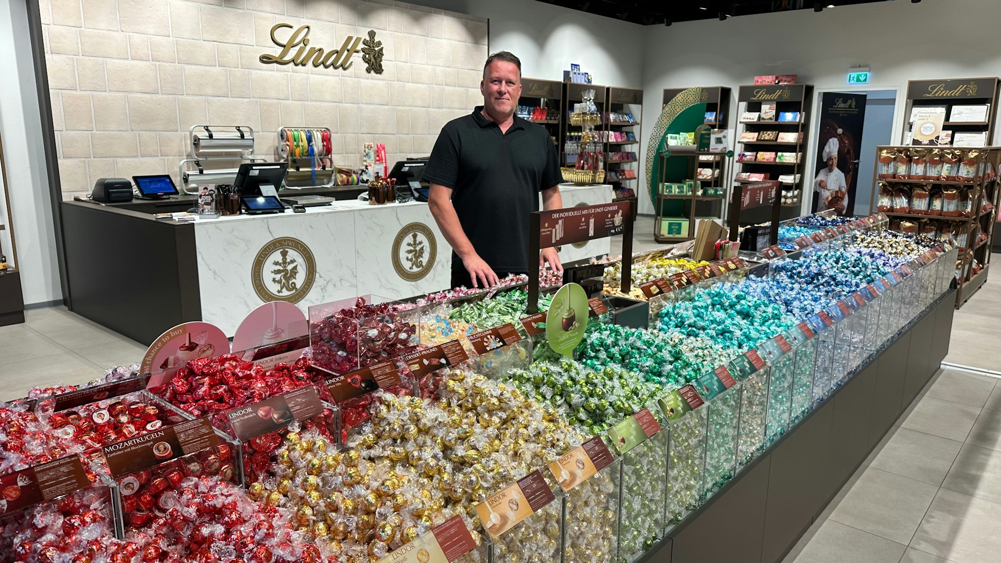 Storemanager Sascha Leeser in seinem Lindt-Shop. In den bunten Farben des Regenbogens sind süßen Versuchungen in der Ladenmitte angeordnet.