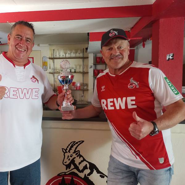 Zwei Fußballfans stehen mit einem Pokal in einem rot-weiß geschmückten Raum.