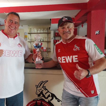 Zwei Fußballfans stehen mit einem Pokal in einem rot-weiß geschmückten Raum.
