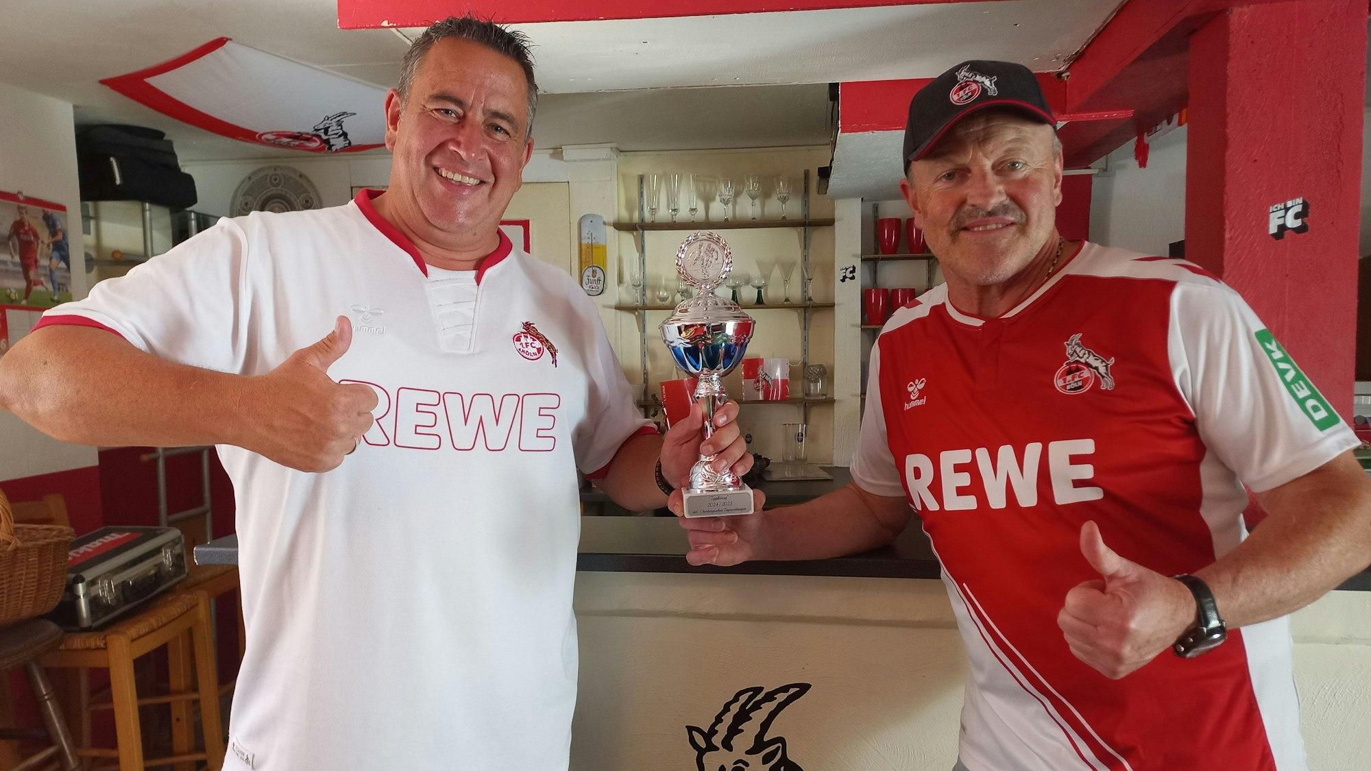 Zwei Fußballfans stehen mit einem Pokal in einem rot-weiß geschmückten Raum.