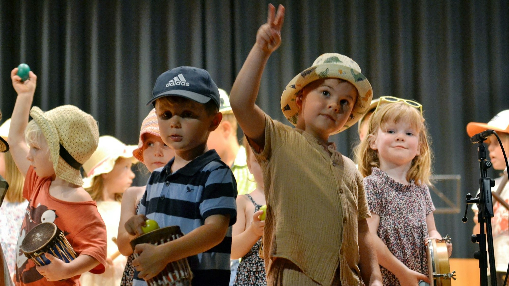 Eine Gruppe kleiner Kinder steht auf einer Bühne, einige haben Trommeln in der Hand, ein Kind mit Hut zeigt ein Peace-Zeichen.