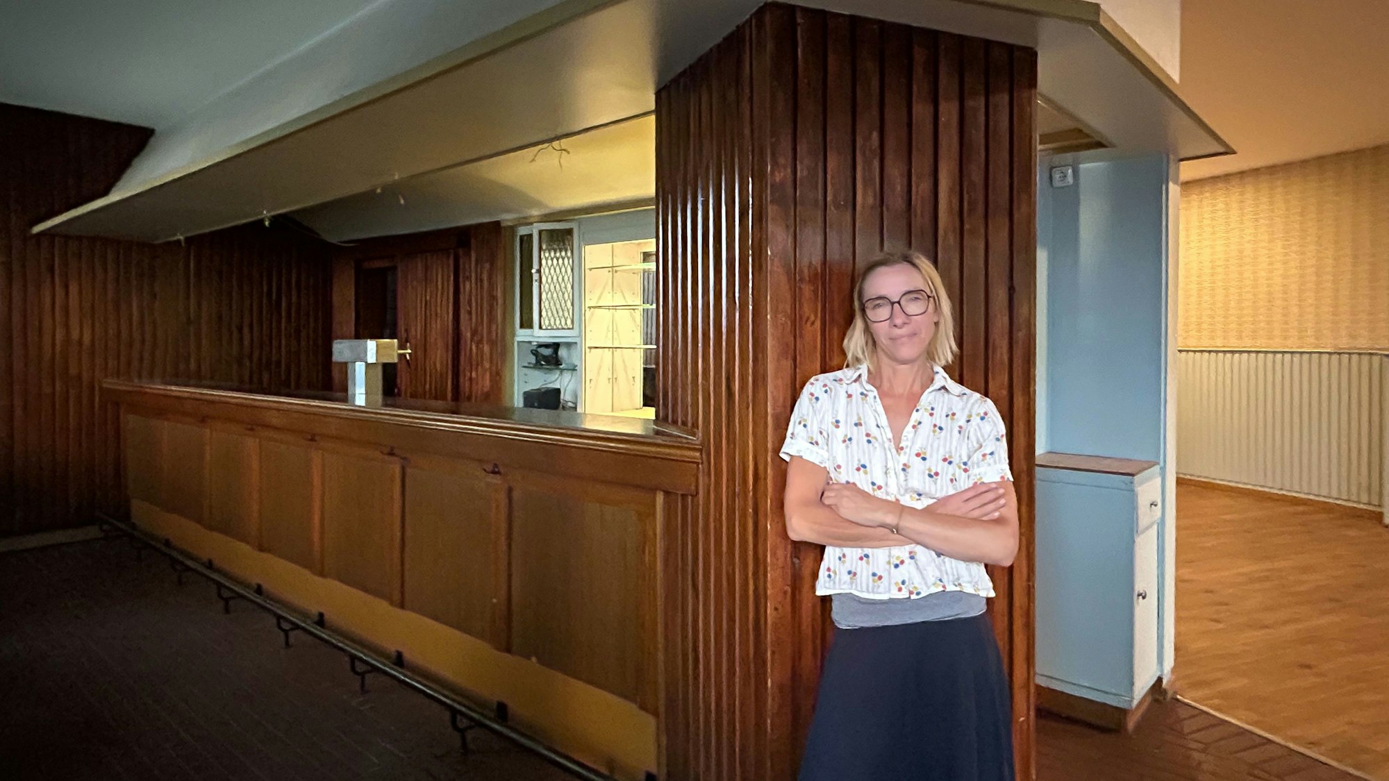 Eine blonde Frau mit Brille steht mit verschränkten Armen an der Seite der Theke ihrer leergeräumten Kneipe Helmholtz.