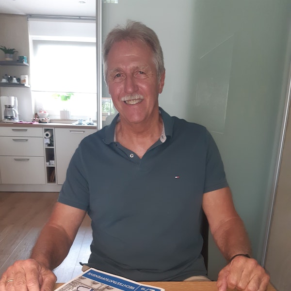 Der Richter hat graue Haare und einen Schnurbart. Er trägt ein graugrünes Polohemd.