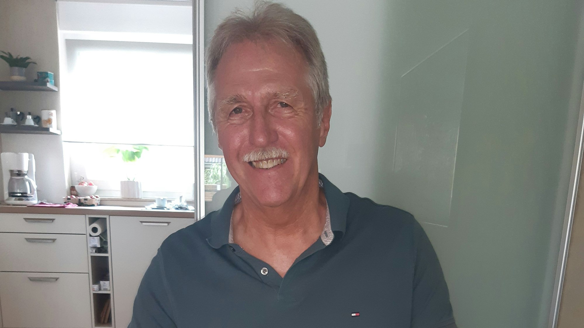 Der Richter hat graue Haare und einen Schnurbart. Er trägt ein graugrünes Polohemd.