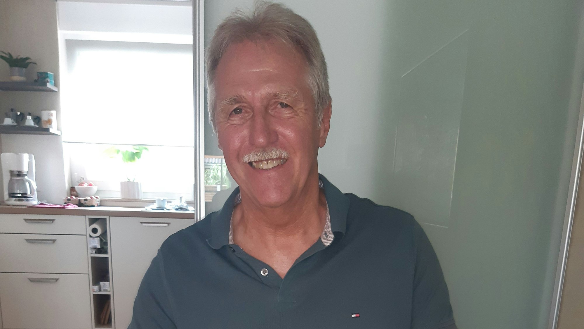 Der Richter hat graue Haare und einen Schnurbart. Er trägt ein graugrünes Polohemd.