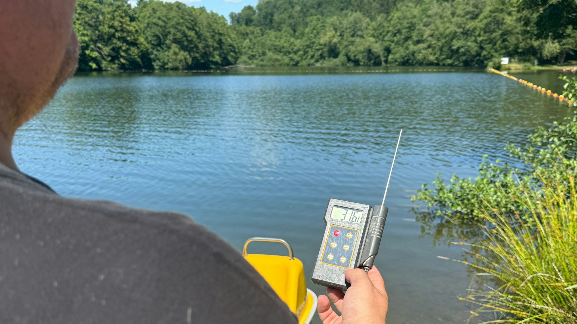 Ein Mann hält an einem Badesee ein Thermometer in der Hand, das 31,6 Grad anzeigt.