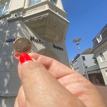 Zu sehen ist eine Ein-Euro-Münze und im Hintergrund das Brühler Rathaus.