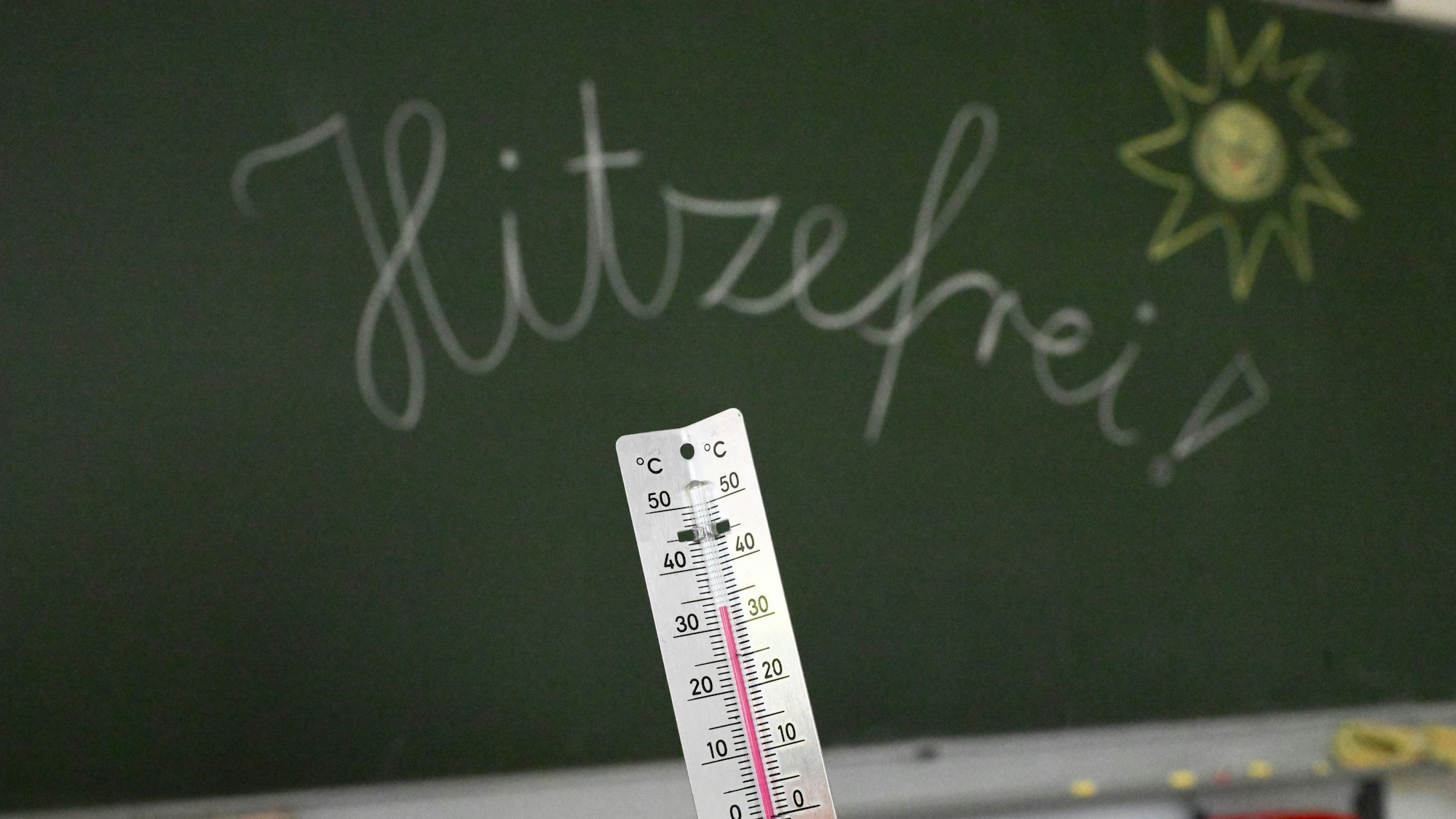 33 Grad werden auf einem Thermometer in einem Klassenzimmer einer Grundschule angezeigt. Auf einer Tafel im Hintergrund hat eine Lehrerin das Wort «Hitzefrei» geschrieben.