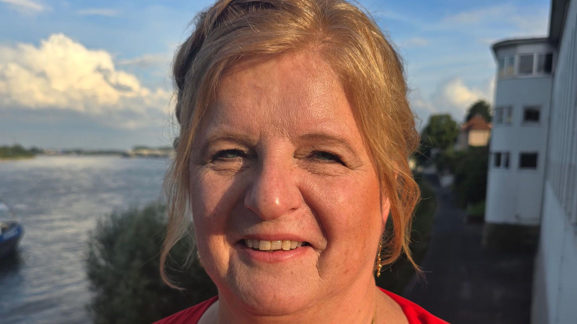 Auf dem Foto ist eine Frau mit blondem Haar zu sehen. Hinter ist fließt der Rhein.