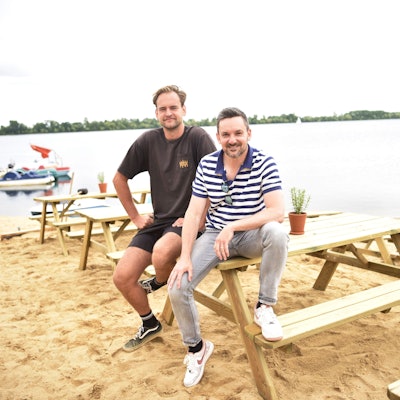 Lukas Funck (links) und Björn Blindert von der surft.GmbH sitzen auf neuen Sitzmöbeln am neuen surft.beach am Zülpicher Wassersportsee. Im Hintergrund sind der See sowie einige Boote zu sehen.