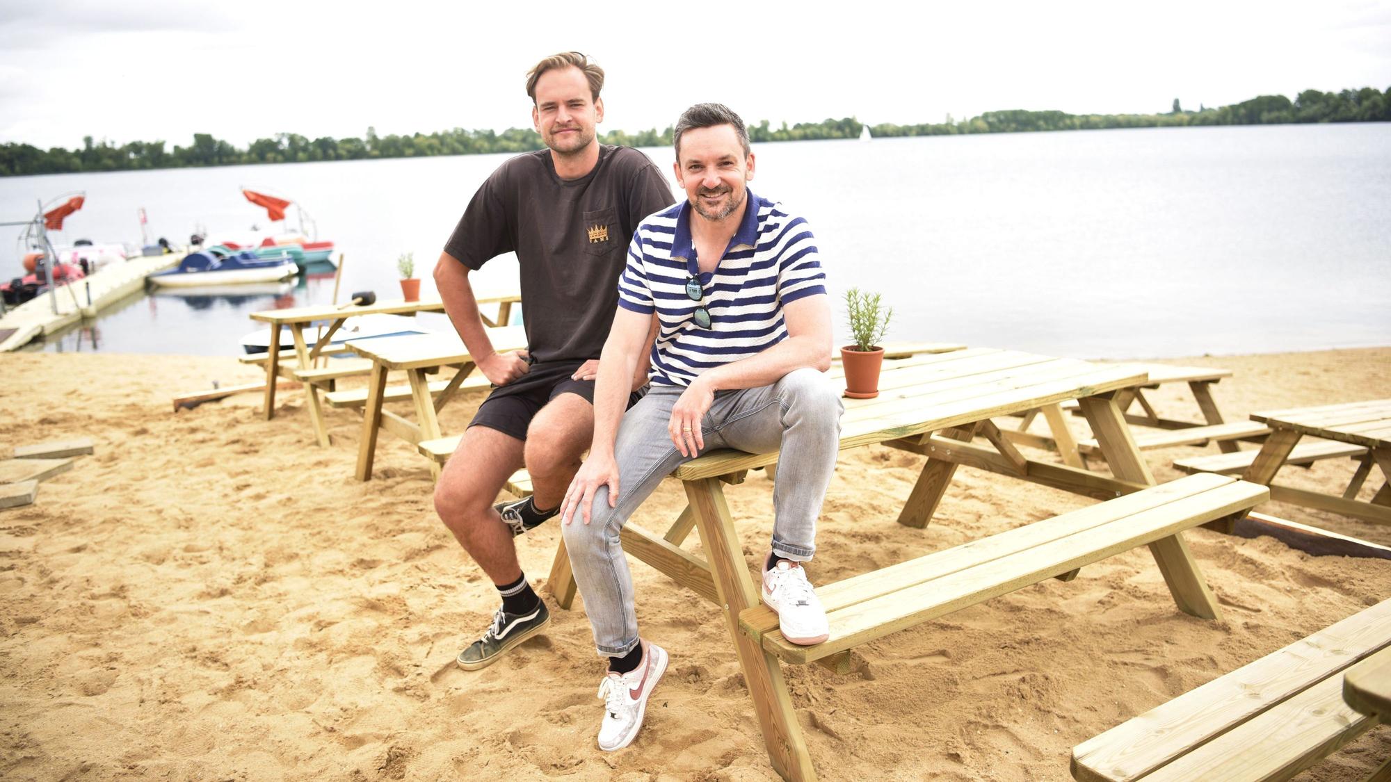 Lukas Funck (links) und Björn Blindert von der surft.GmbH sitzen auf neuen Sitzmöbeln am neuen surft.beach am Zülpicher Wassersportsee. Im Hintergrund sind der See sowie einige Boote zu sehen.