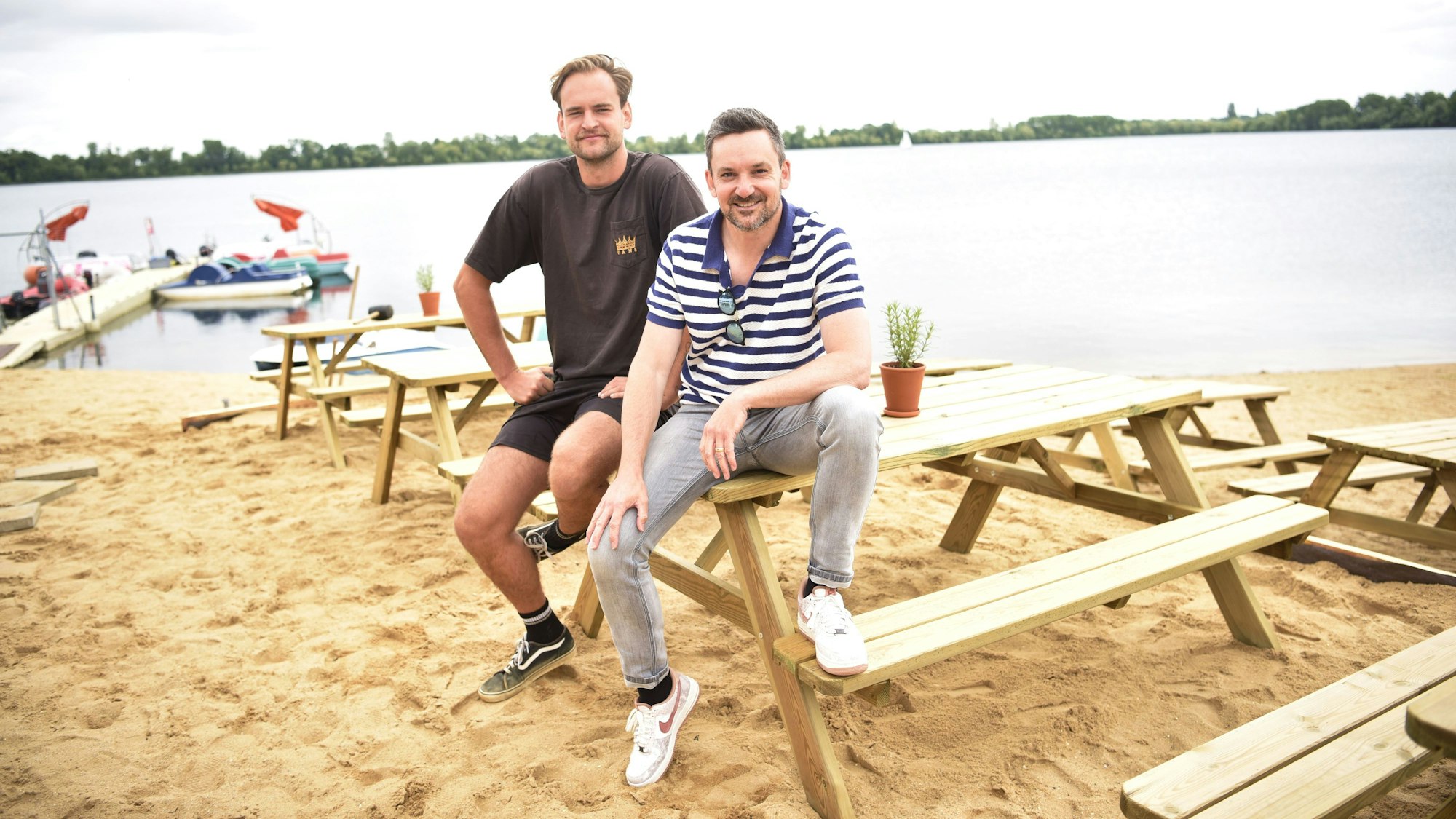 Lukas Funck (links) und Björn Blindert von der surft.GmbH sitzen auf neuen Sitzmöbeln am neuen surft.beach am Zülpicher Wassersportsee. Im Hintergrund sind der See sowie einige Boote zu sehen.