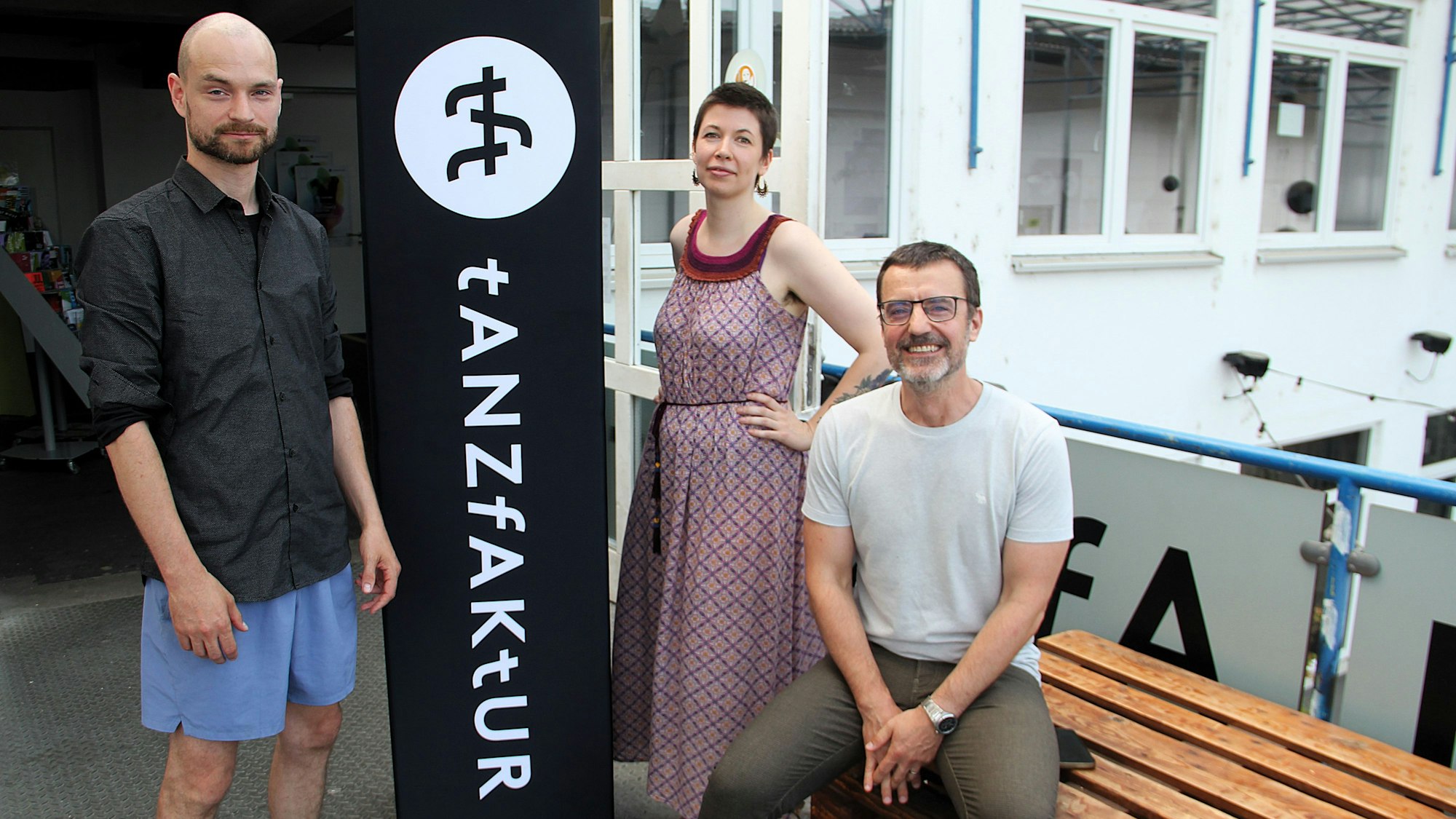 Zwei Männer und eine Frauen stehen bzw. sitzen neben einem Säulenbanner auf dem TanzFaktur steht.