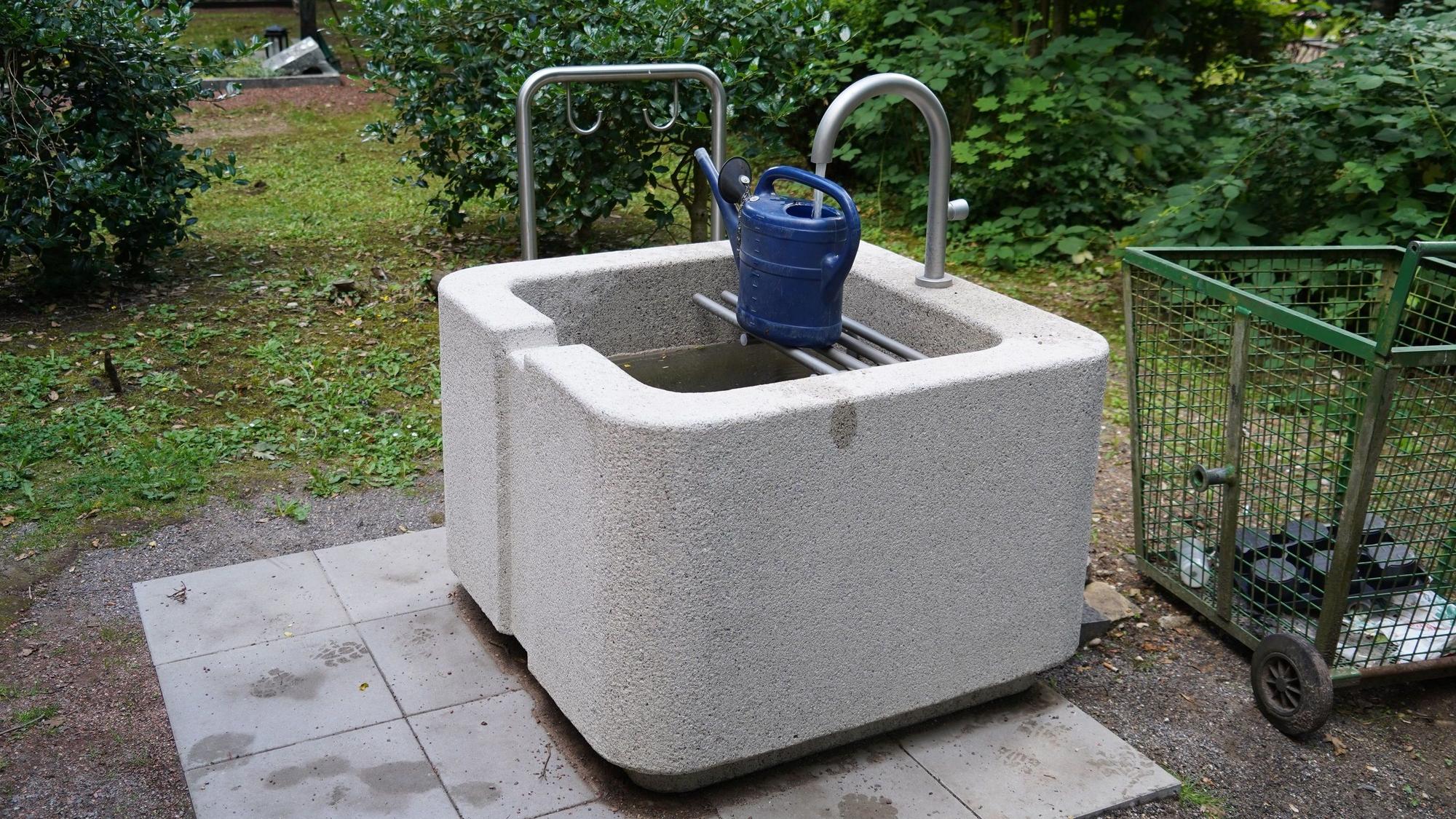 Auf dem Bild ist ein graues Stein-Wasserbecken mit einer blauen Gießkanne zu sehen.