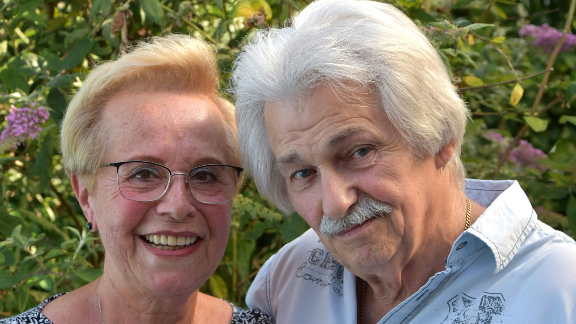 Das Bild zeigt die beiden in einer Nahaufnahme, Köpfe leicht zueinander gelehnt. Hinter ihnen Büsche und ein paar Blüten.