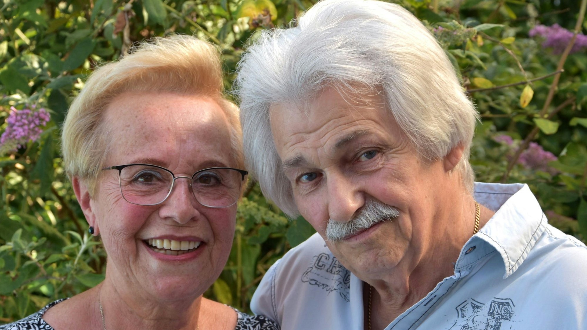 Das Bild zeigt die beiden in einer Nahaufnahme, Köpfe leicht zueinander gelehnt. Hinter ihnen Büsche und ein paar Blüten.