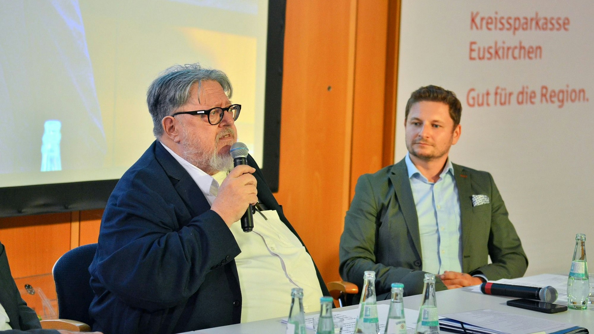 Bernd Kolvenbach und George Tulbure sitzen in Anzügen an einem Tisch, auf dem Wasserflaschen stehen. Bernd Kolvenbach trägt eine Brille und spricht in ein Mikrofon.