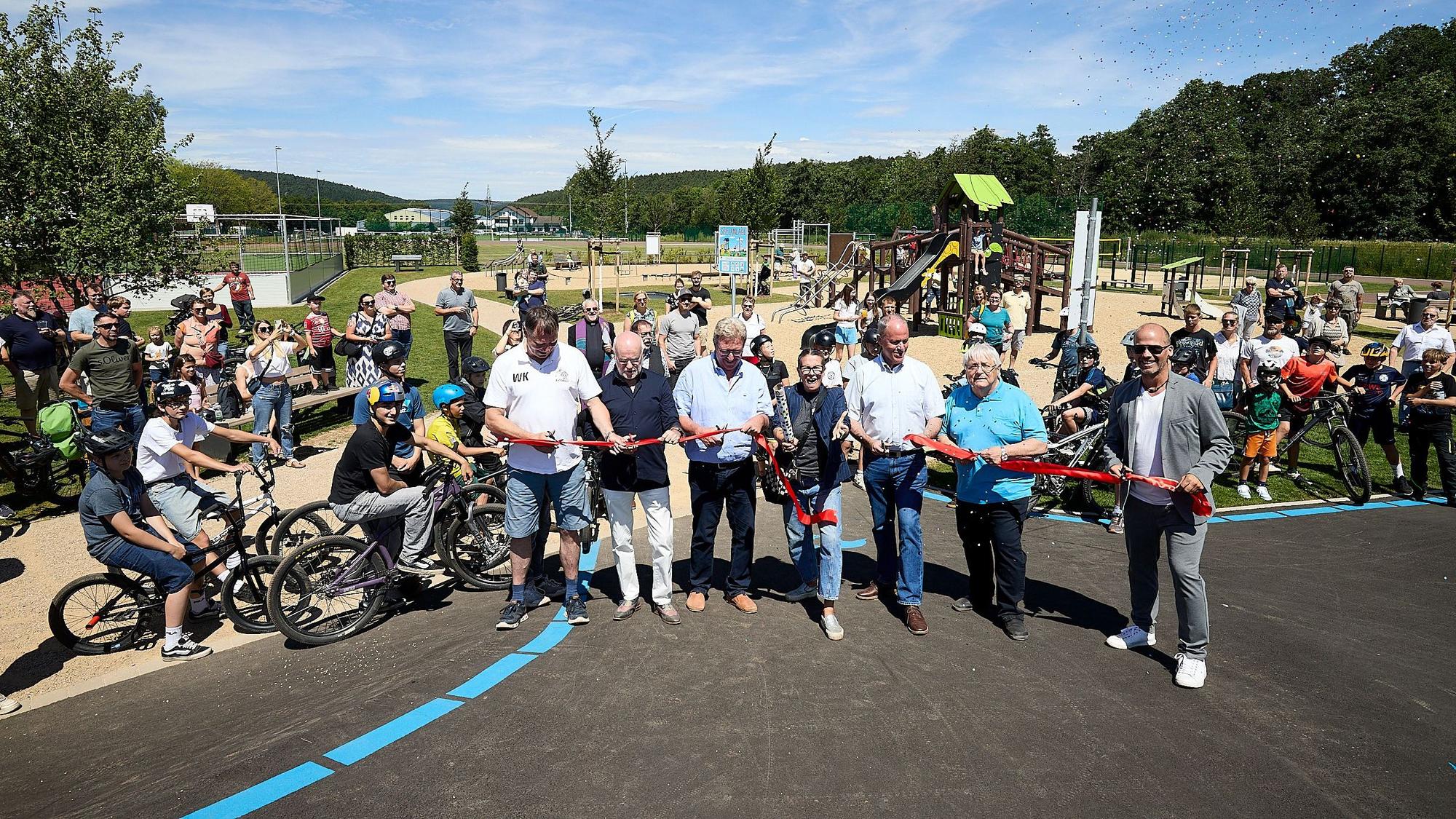 Eine Gruppe Menschen steht auf einer asphaltierten Pump-Track-Bahn und schneidet ein rotes Band durch. Im Hintergrund sind Zuschauer und Sport- und Spielgeräte zu erkennen.