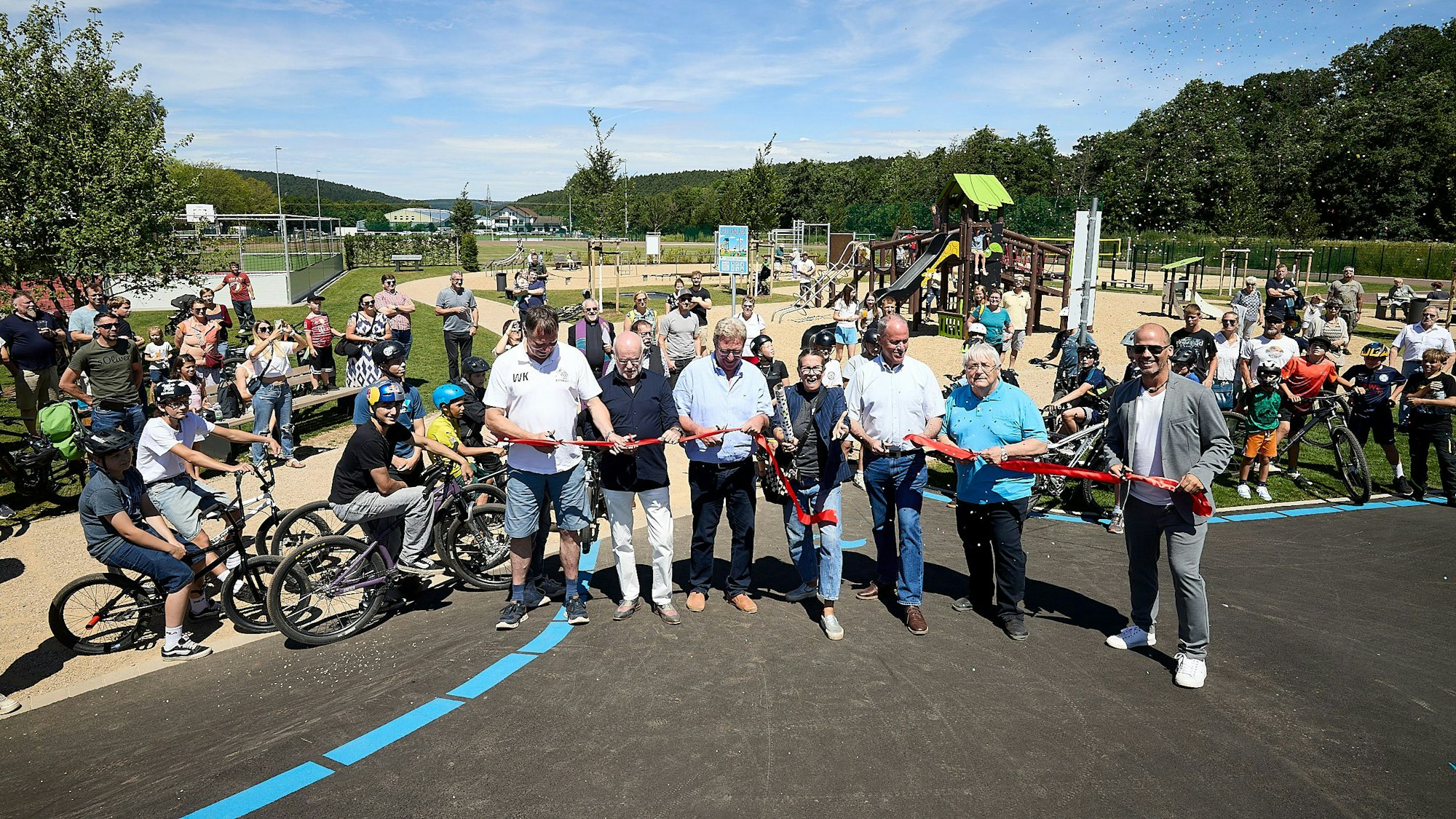 Eine Gruppe Menschen steht auf einer asphaltierten Pump-Track-Bahn und schneidet ein rotes Band durch. Im Hintergrund sind Zuschauer und Sport- und Spielgeräte zu erkennen.