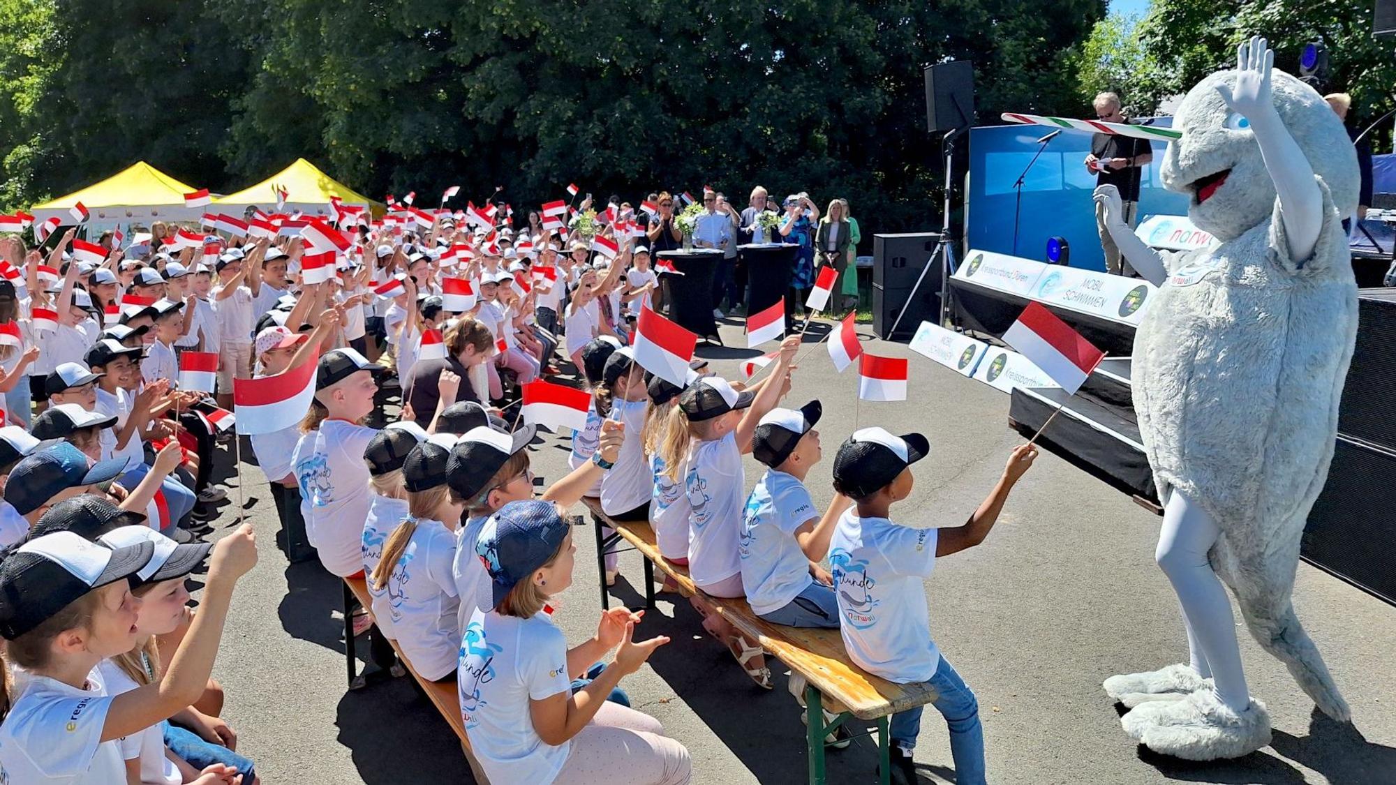 Kinder in hellen T-Shirts sitzen auf Bierbänken vor einer Bühne und schwenken rot-weiße Fahnen. Vor der Bühne steht ein als Narwal verkleidetes Maskottchen.