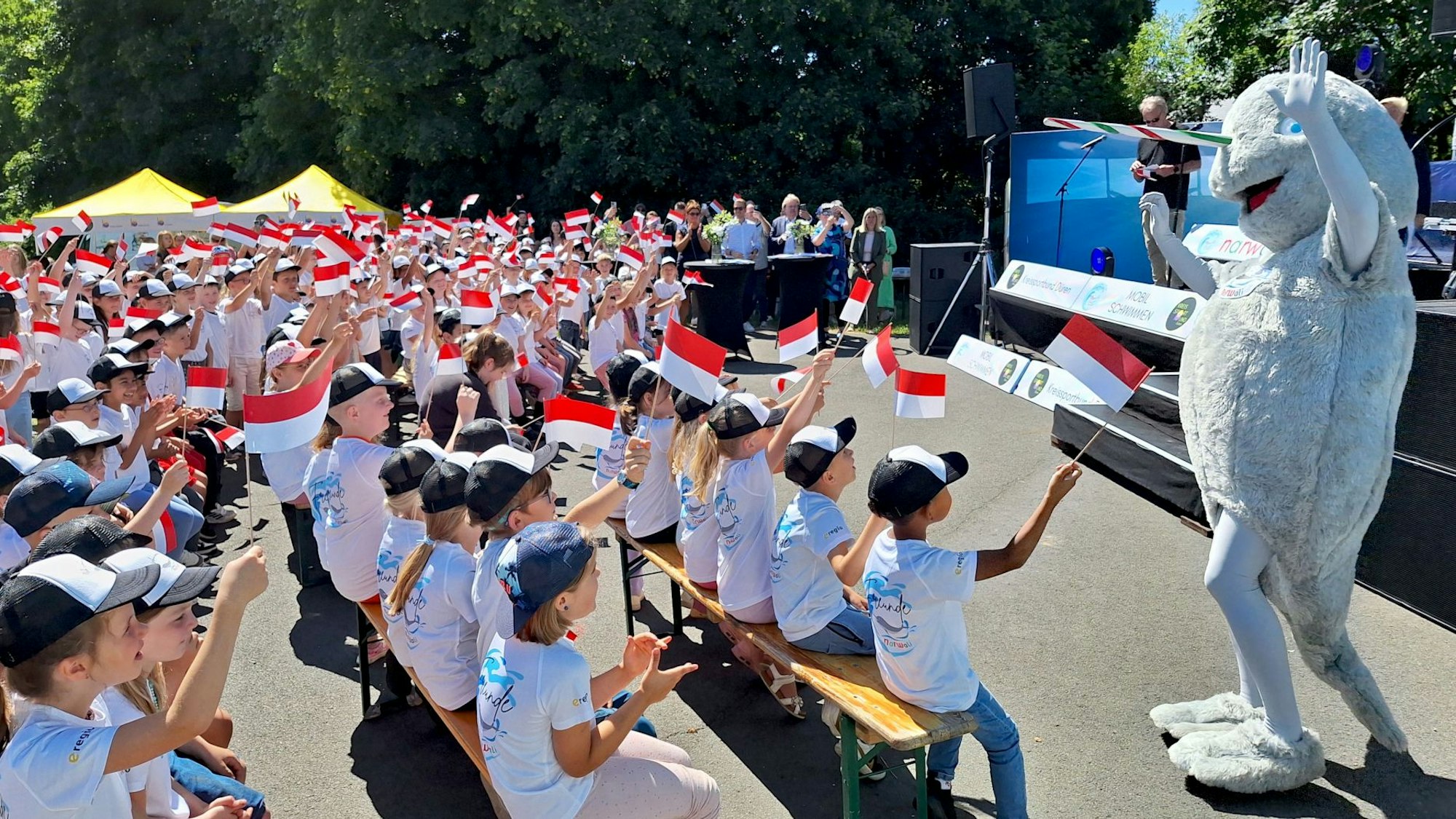 Kinder in hellen T-Shirts sitzen auf Bierbänken vor einer Bühne und schwenken rot-weiße Fahnen. Vor der Bühne steht ein als Narwal verkleidetes Maskottchen.