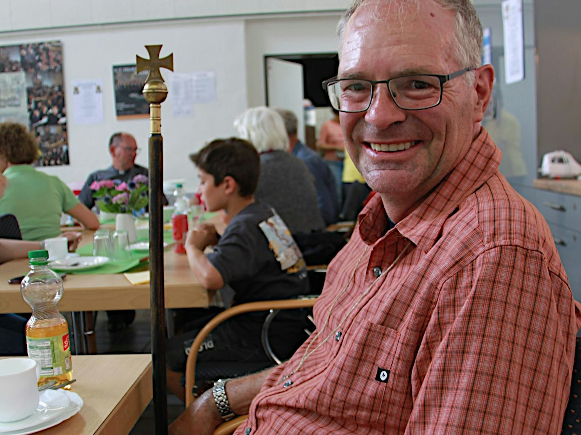 Ein Mann in rot-kariertem Freizeithemd sitzt an einem Tisch und lacht in die Kamera.