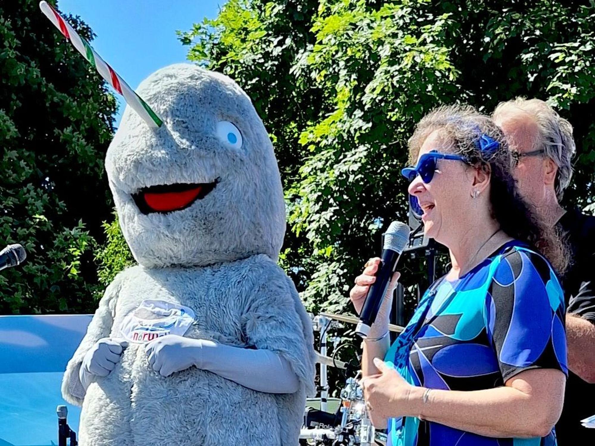 Eine Frau in blau-gemustertem Kleid steht neben einem Narwal-Maskottchen und spricht in ein Mikrofon.