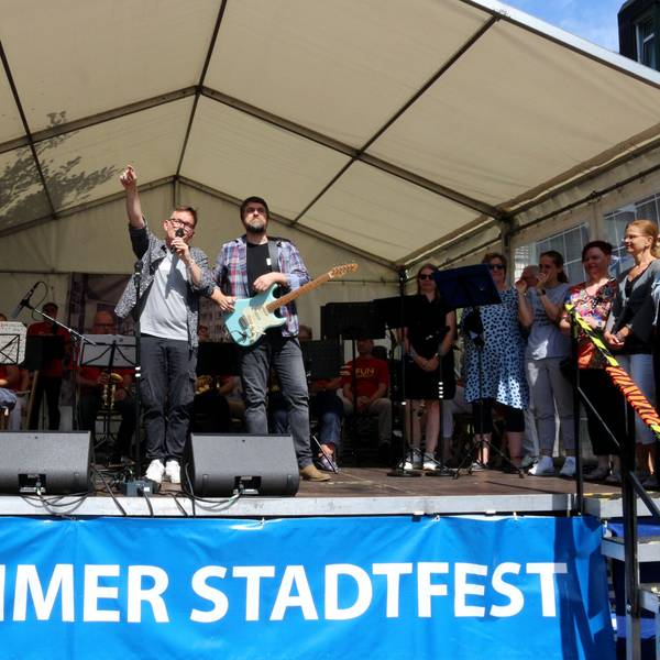 Auf der Bühne: Ein Musiker am Keyboard, einer an der Gitarre, einer mit einem Mikro, an der Seite rechts stehen mehrere Sängerinnen.