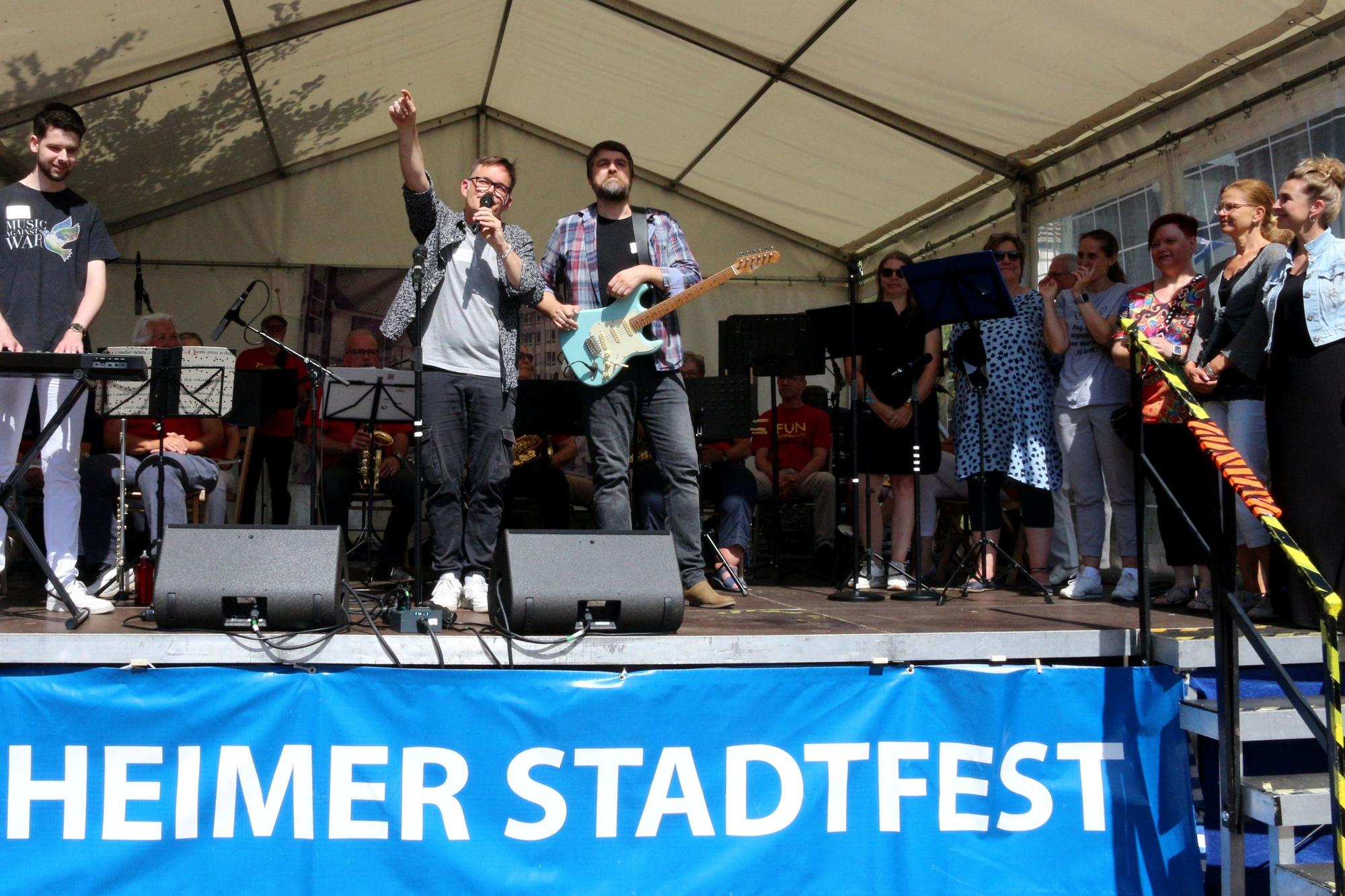 Auf der Bühne: Ein Musiker am Keyboard, einer an der Gitarre, einer mit einem Mikro, an der Seite rechts stehen mehrere Sängerinnen.