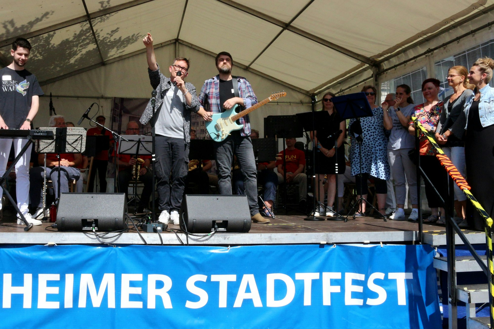 Auf der Bühne: Ein Musiker am Keyboard, einer an der Gitarre, einer mit einem Mikro, an der Seite rechts stehen mehrere Sängerinnen.