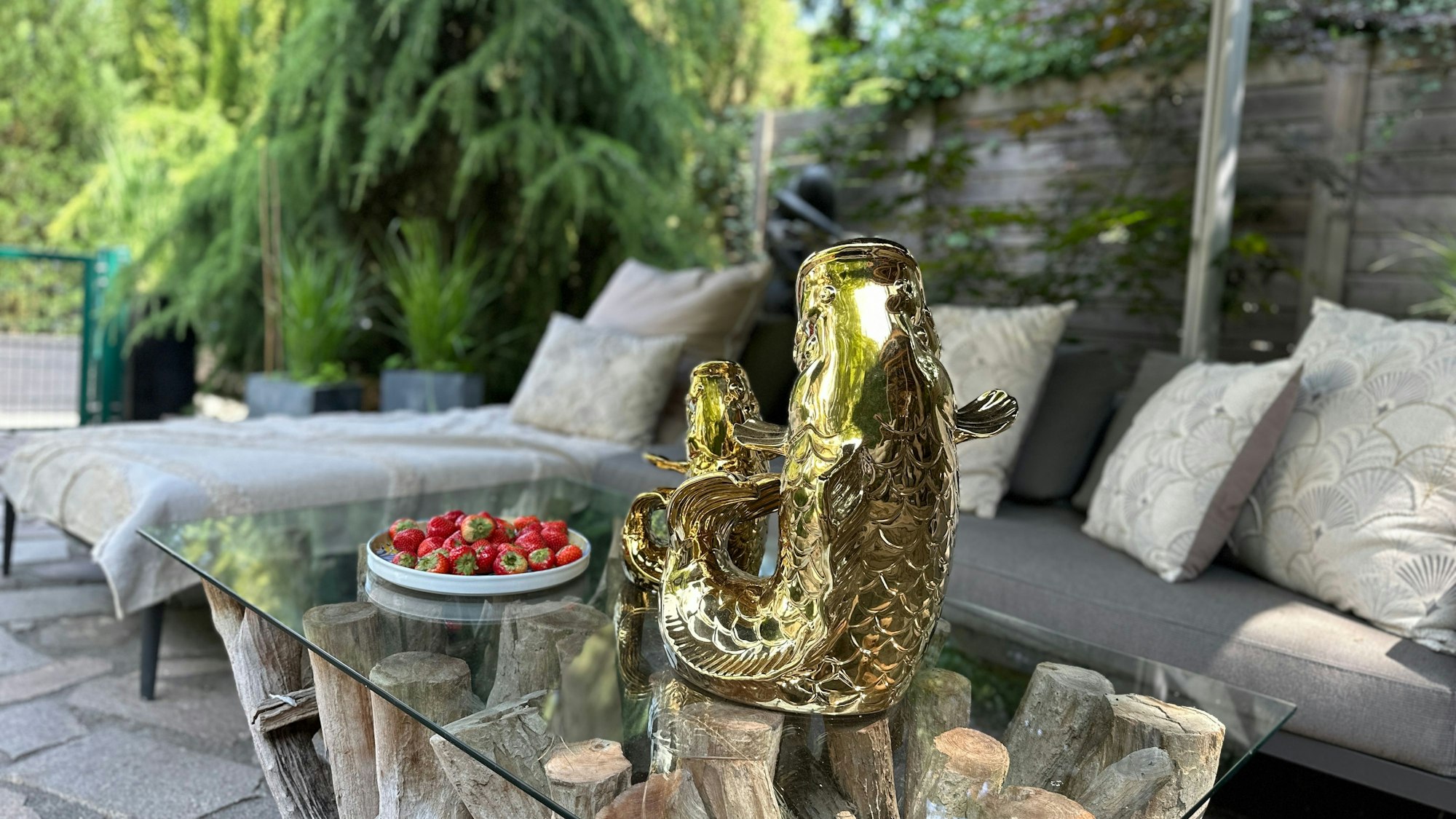 Auf einer Steinterrasse steht ein Glastisch mit einer goldfarbenen Fisch-Wasserkaraffe.