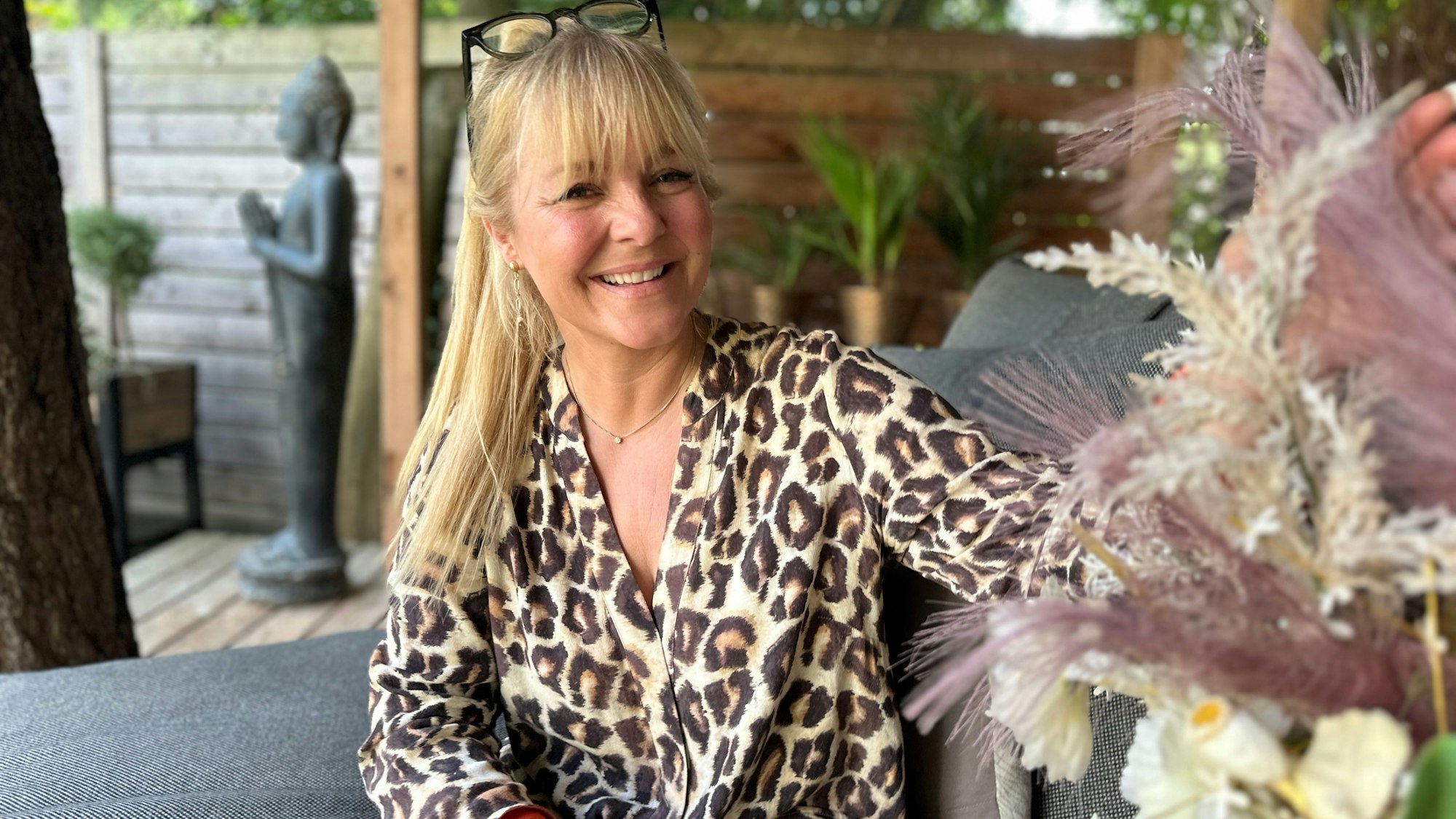 Eine Frau mit blondem Pferdeschwanz und in Leoprint-Bluse sitzt auf einer Terrasse, die geschmackvoll dekoriert ist.