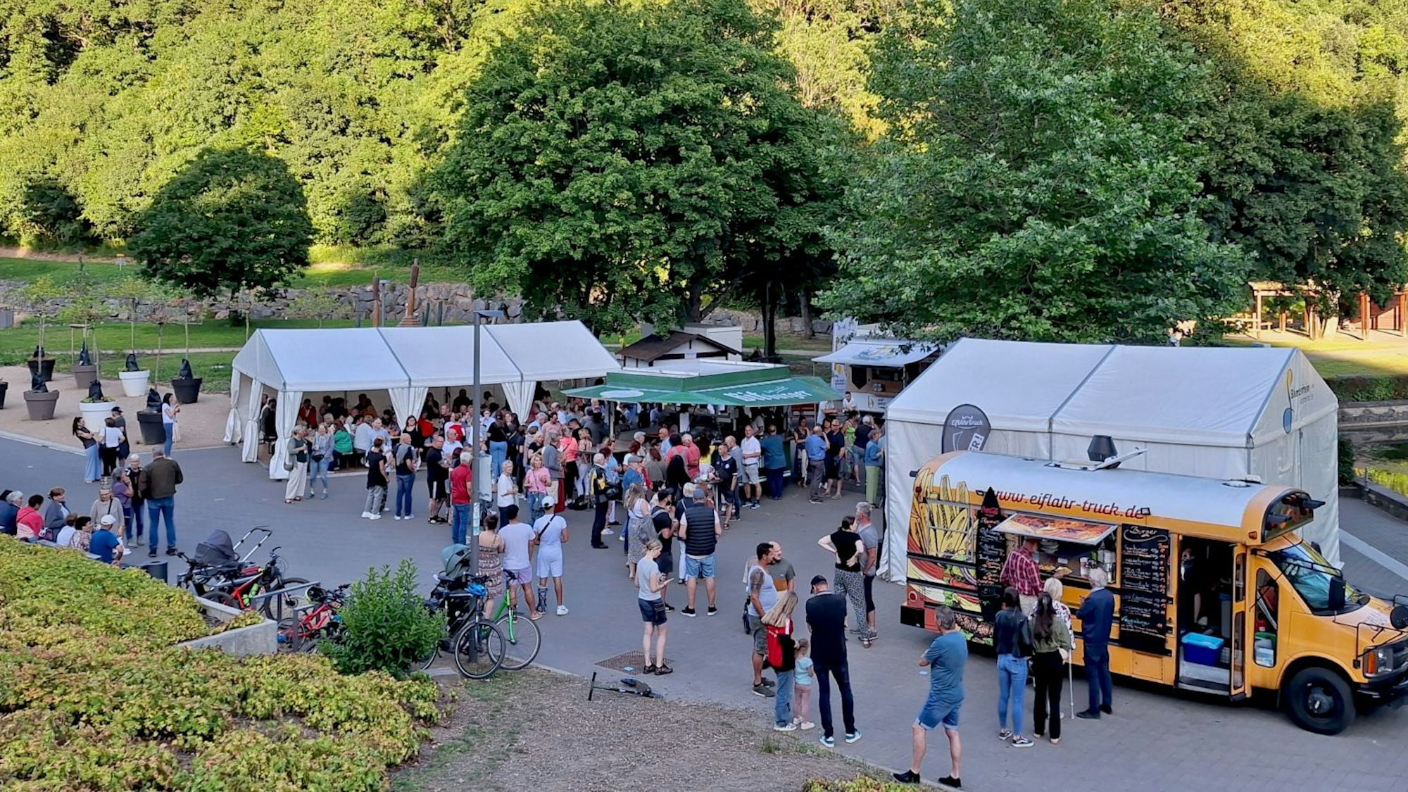Eine erhöhte Seitenansicht zeigt den Platz, auf dem das Festival "Sommer am See" in Blankenheim stattfindet. Rechts steht ein gelber Foodtruck, links dahinter befinden sich Zelte, viele Menschen tummeln sich auf dem Platz.