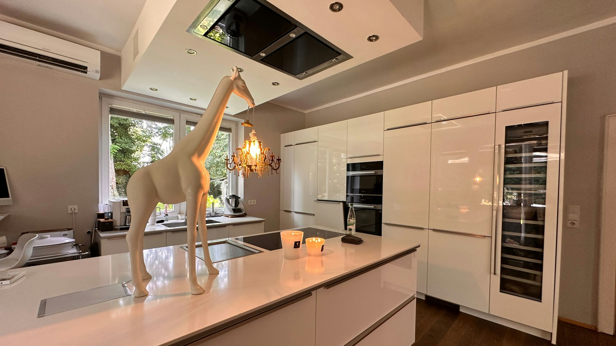 In einer geräumigen weißen Küche steht eine weiße Lampe in Form einer Giraffe auf dem Tresen, der auch das Spülbecken enthält.