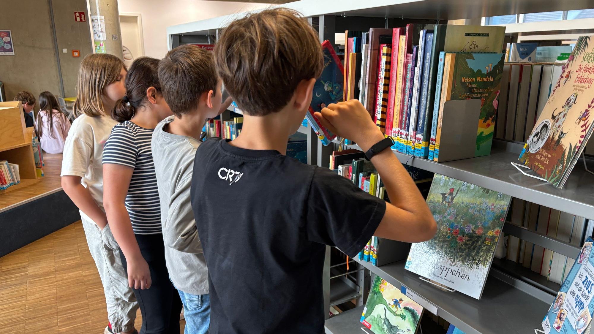 Vier Kinder stehen an einem Bücherregal. Ein Junge nimmt ein Buch heraus.