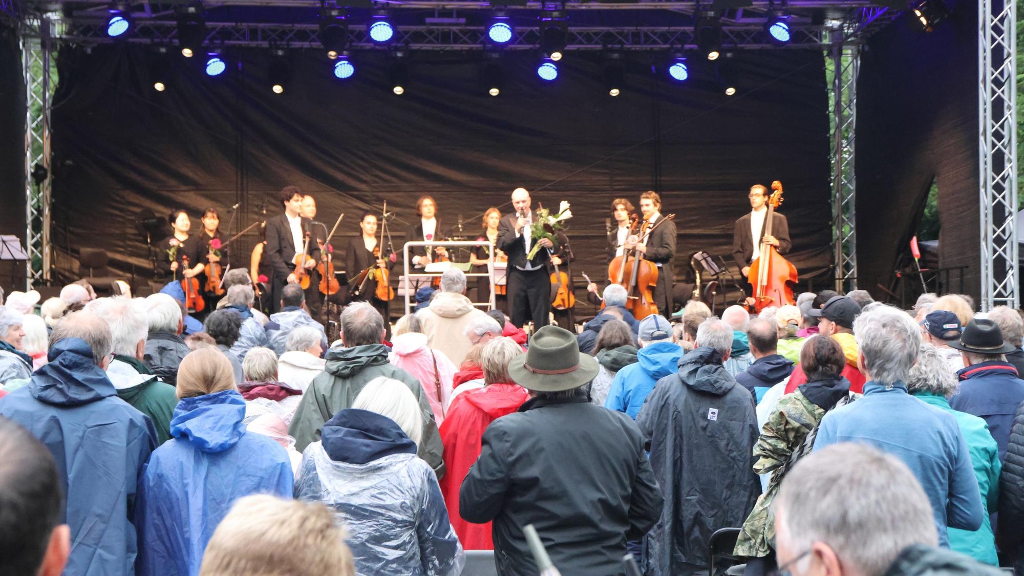 Ein Orchester auf der Bühne, davor Zuhörer in Regencapes.