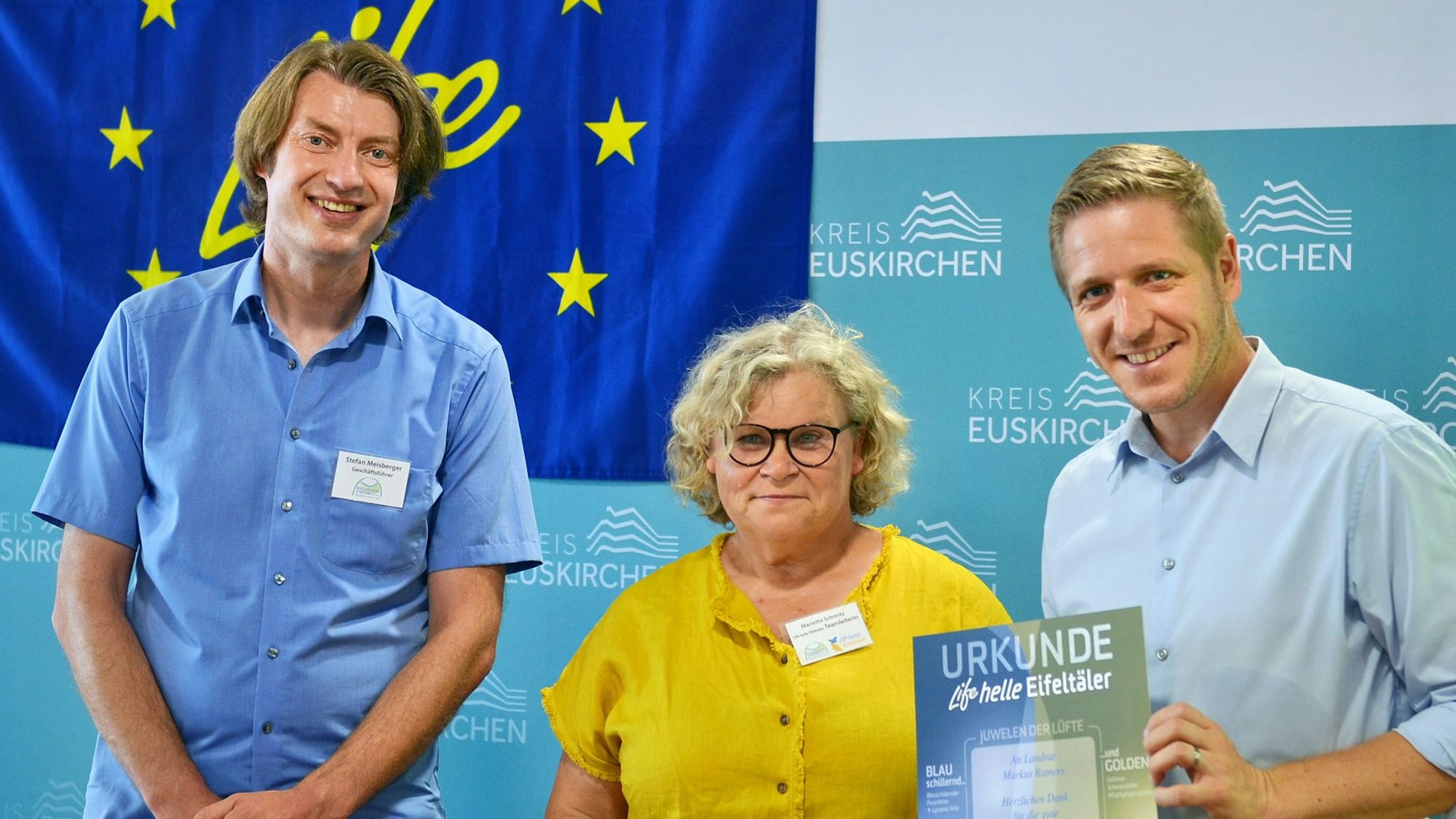 Stefan Meisberger, Marietta Schmitz und Markus Ramers stehen nebeneinander und grinsen in die Kamera. Markus Ramers hält dabei eine Urkunde auf der „Life helle Eifeltäler“ steht.