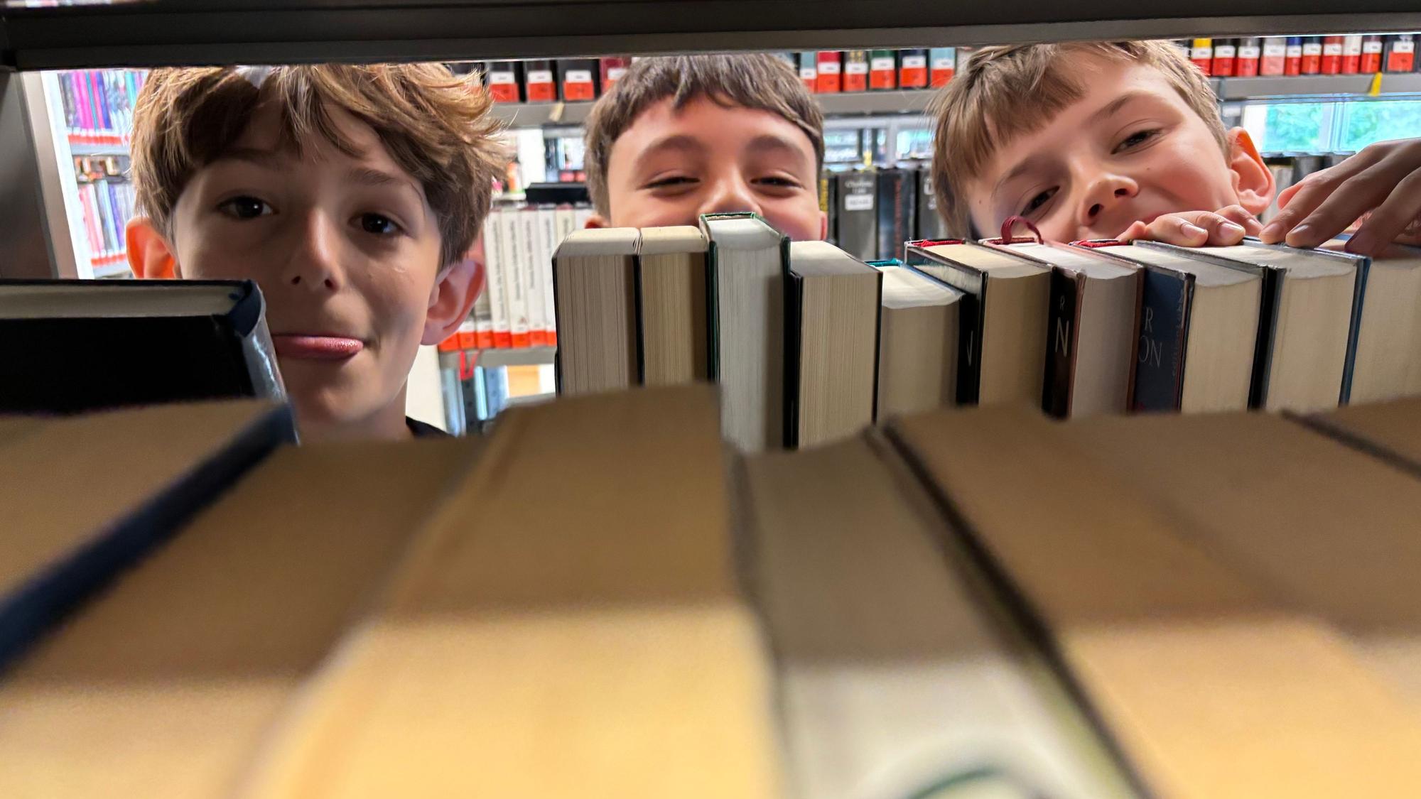 Die Köpfe von drei Jungen schauen hinter Büchern in einem Regal hervor. Einer der drei streckt seine Zunge raus.