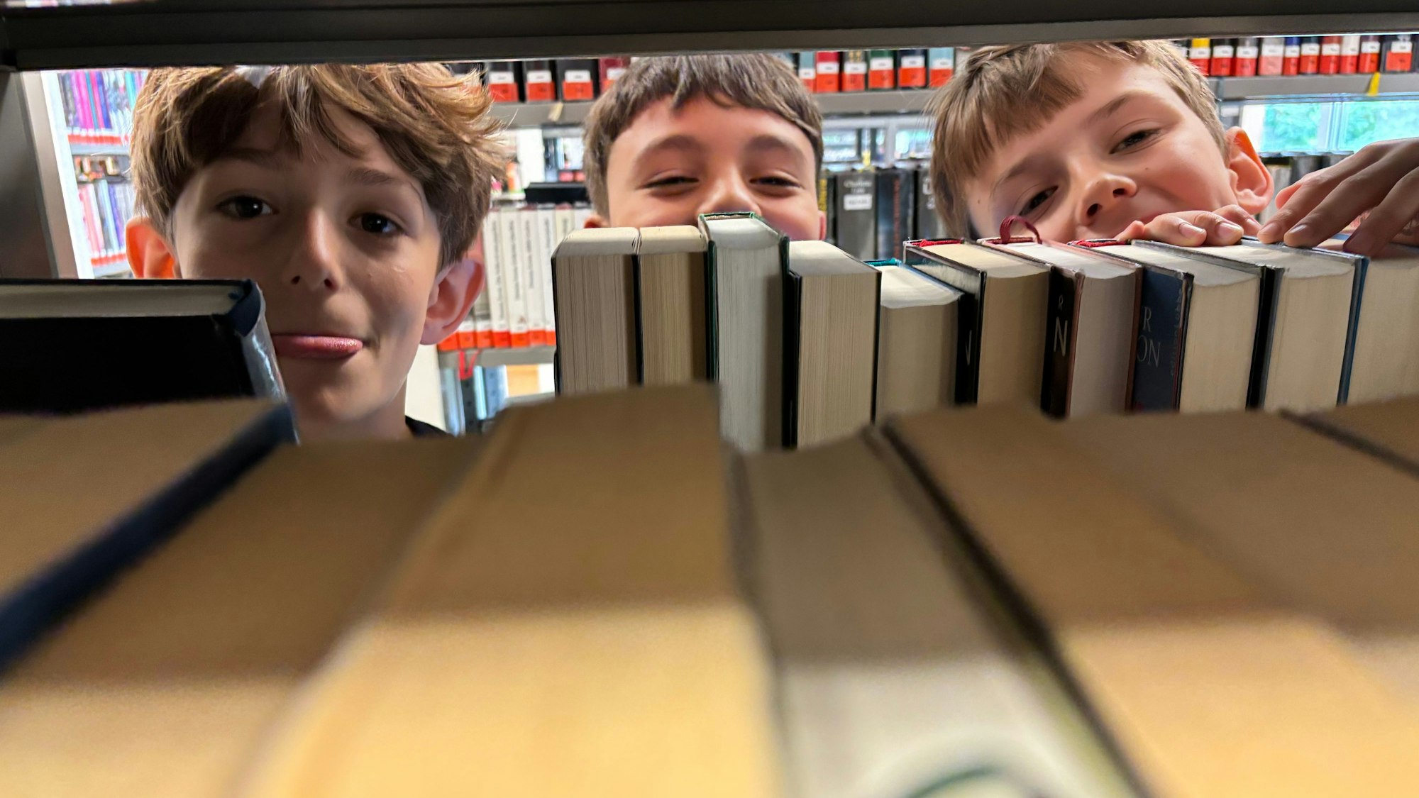Die Köpfe von drei Jungen schauen hinter Büchern in einem Regal hervor. Einer der drei streckt seine Zunge raus.