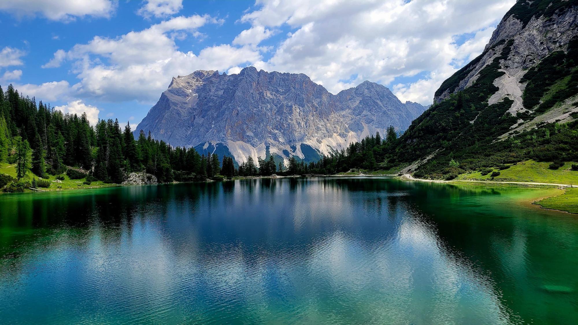Blick auf den türkisblauen, von Bäumen eingerahmten Seebensee, auf dem Wanderweg zur Zugspitze. Im Hintergrund sind Berge zu sehen.
