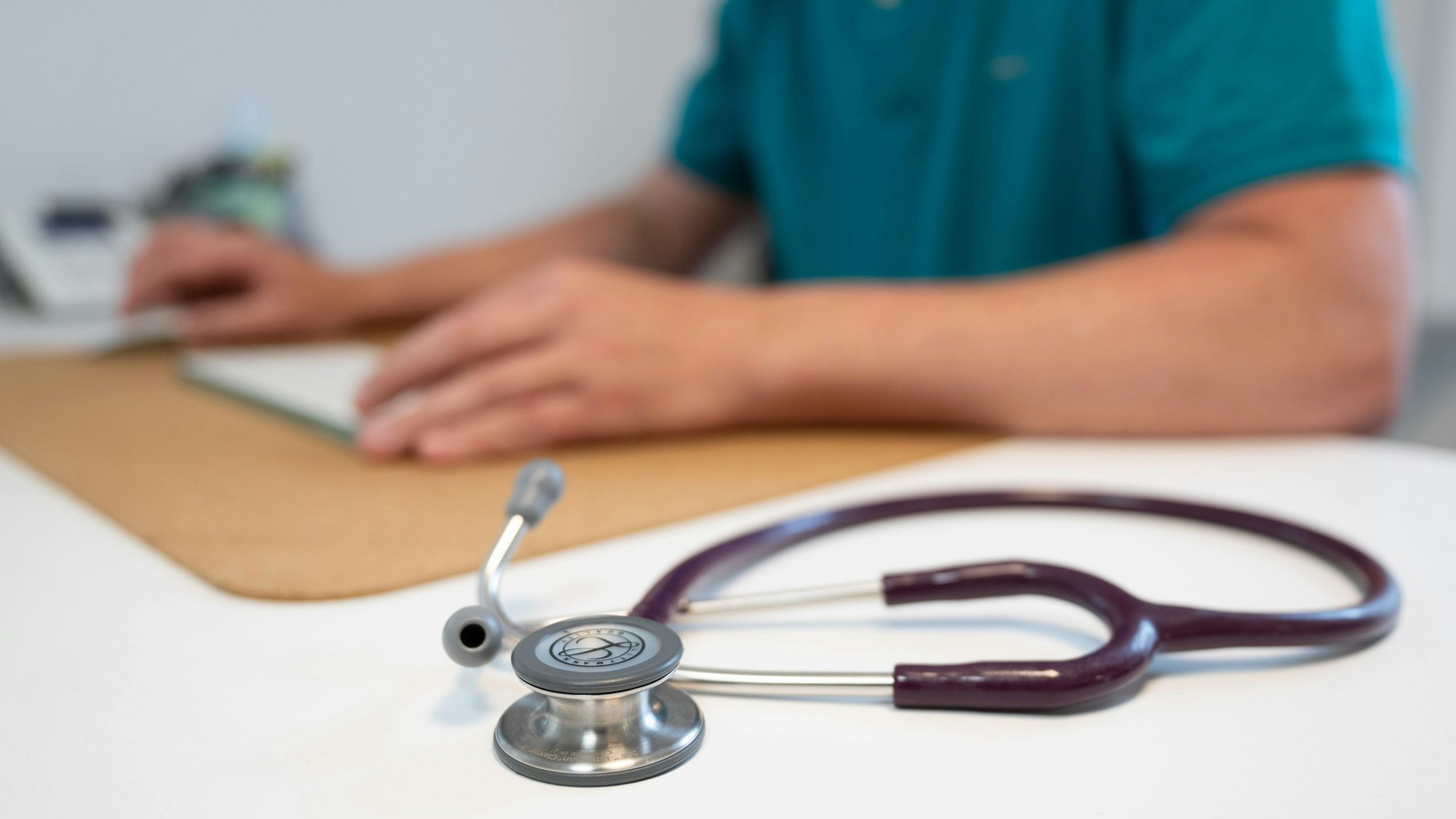 Ein Hausarzt sitzt in einem Sprechzimmer seiner Hausarztpraxis an einem Schreibtisch neben einem Stethoskop und arbeitet am Computer.