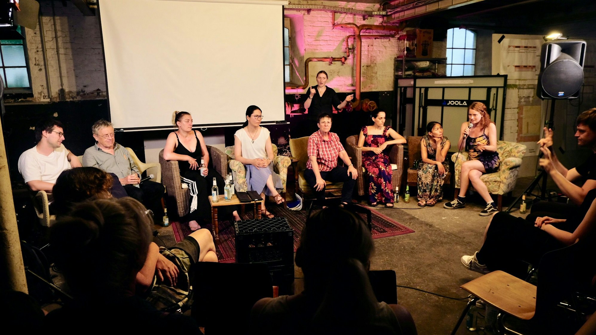 Auf Sesseln in einem großen Raum sitzen Menschen, eine Frau spricht in ein Mikrofon.