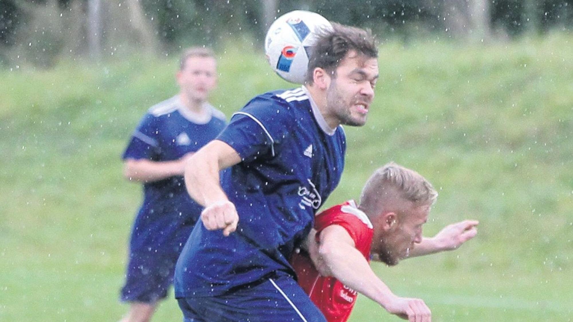 Es regnet auf einem Fußballplatz. Zwei Spieler sind im Zweikampf. Einer der beiden bekommt den Ball gegen den Hinterkopf und hat die Augen geschlossen.