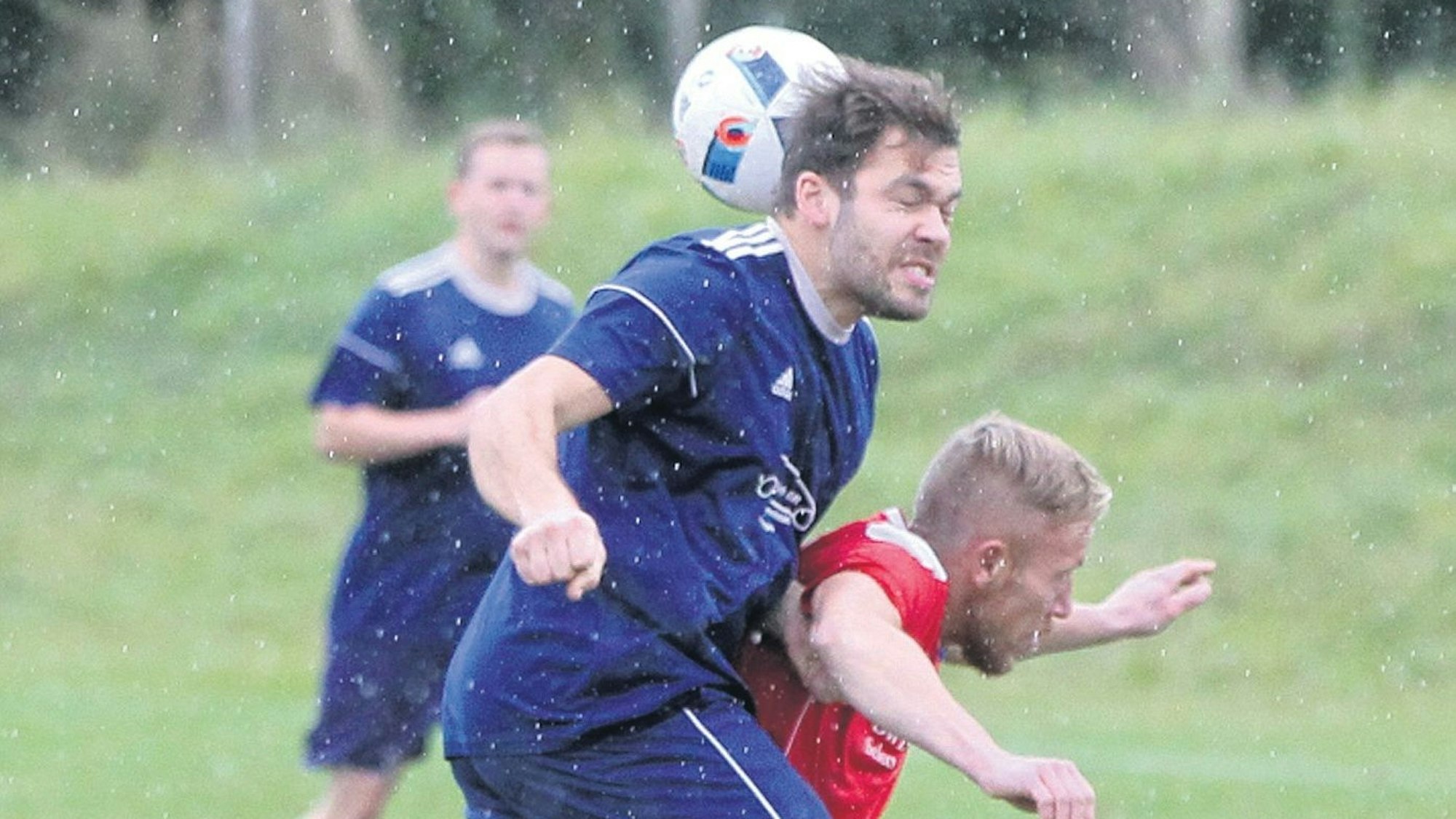 Es regnet auf einem Fußballplatz. Zwei Spieler sind im Zweikampf. Einer der beiden bekommt den Ball gegen den Hinterkopf und hat die Augen geschlossen.