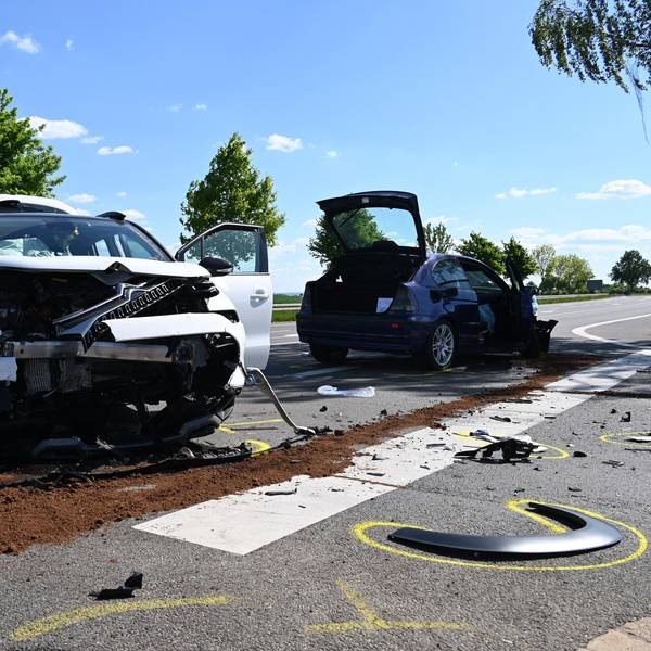 Zu sehen sind zwei Autos nach einem Unfall auf einer Kreuzung, im Hintergrund steht ein Krankenwagen.
