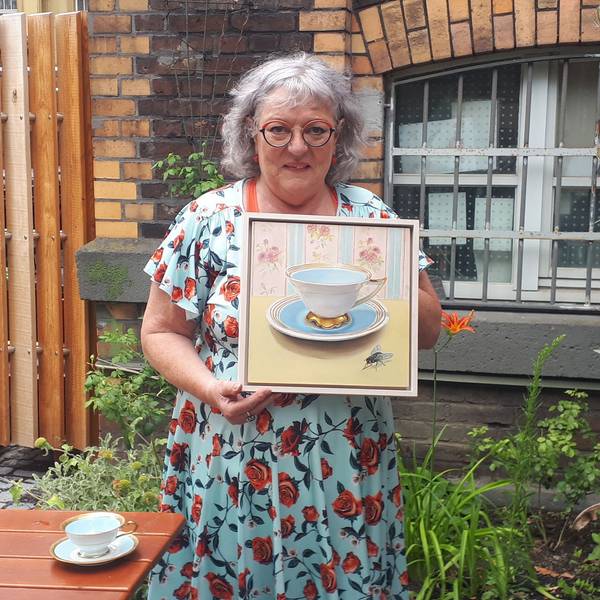 Eine Frau mit grauen Haaren trägt ein hellblaues Kleid mit roten Rosen darauf. Sie steht in einem Garten und hält ein Bild, auf dem eine historische Tasse und eine Fliege zu erkennen sind.