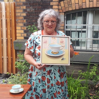 Eine Frau mit grauen Haaren trägt ein hellblaues Kleid mit roten Rosen darauf. Sie steht in einem Garten und hält ein Bild, auf dem eine historische Tasse und eine Fliege zu erkennen sind.