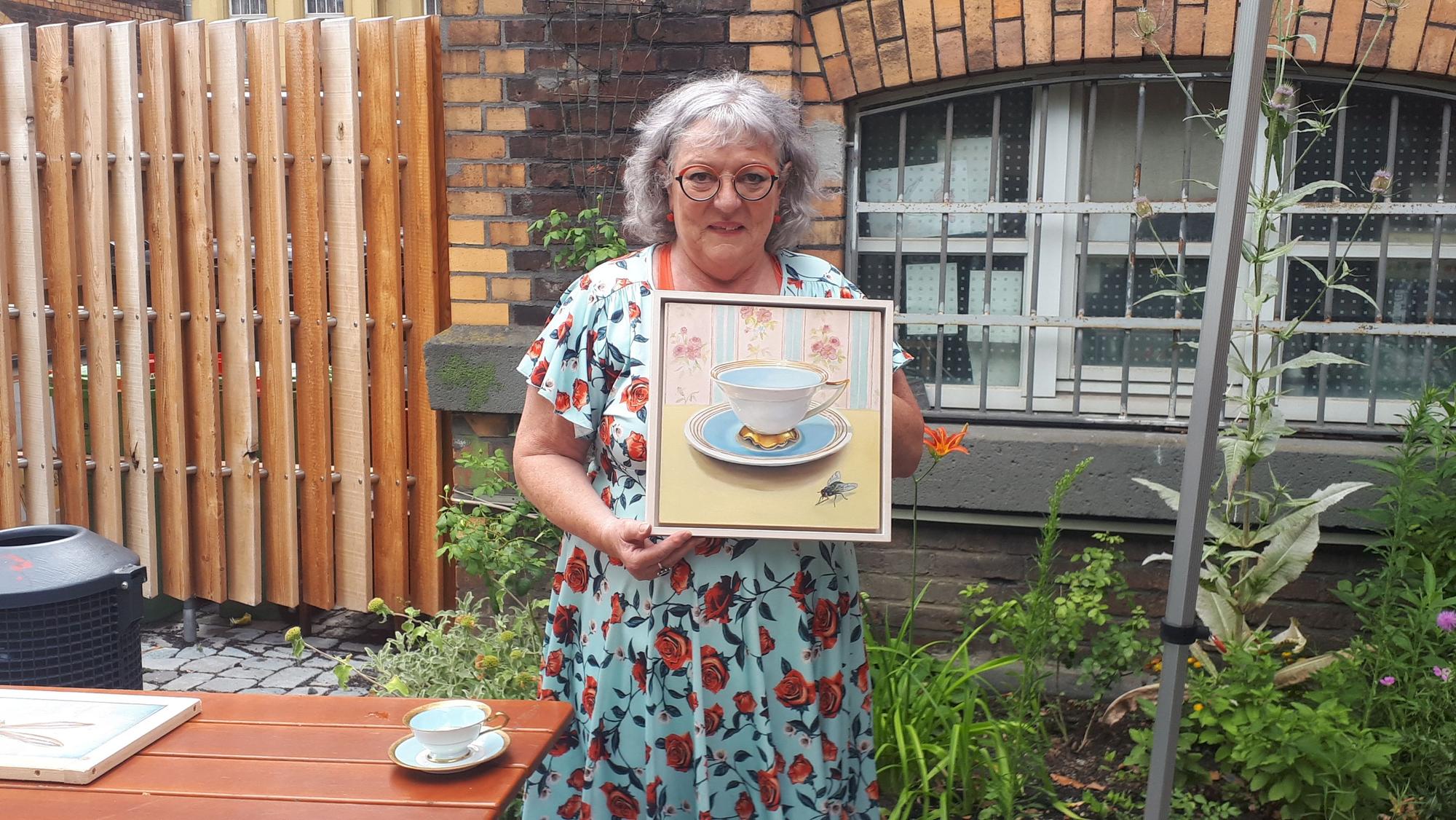 Eine Frau mit grauen Haaren trägt ein hellblaues Kleid mit roten Rosen darauf. Sie steht in einem Garten und hält ein Bild, auf dem eine historische Tasse und eine Fliege zu erkennen sind.